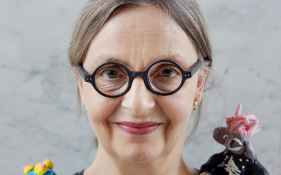 Silmälasipäinen, tummapaitainen nainen, jonka pään päällä ja olkapäillä on pieniä värikkäitä hahmoja