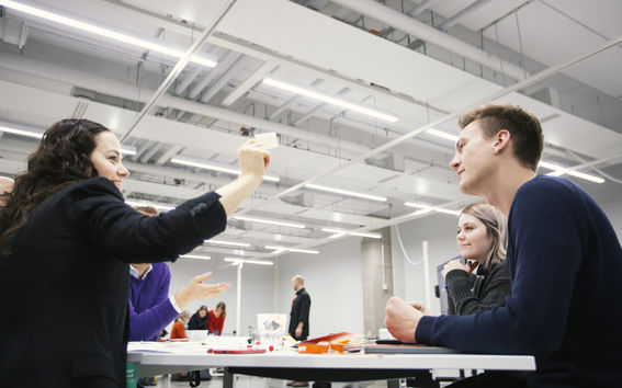 Image of a team around a table working together. One person is holding a post-it up in the air.