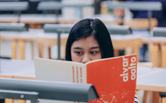 A student's face peaking behind a book while reading in a university library.