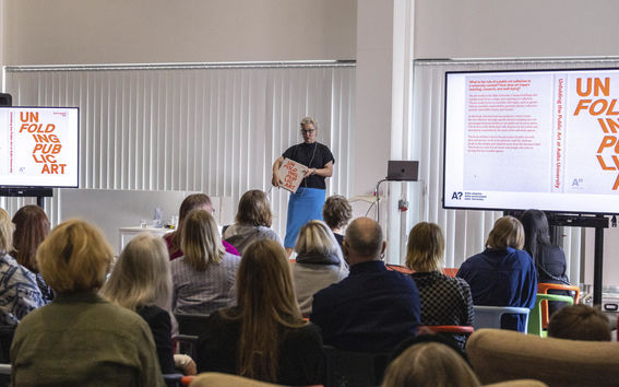 Outi Turpeinen standing on stage talking about Unfolding public art book to a seated audience facing her.