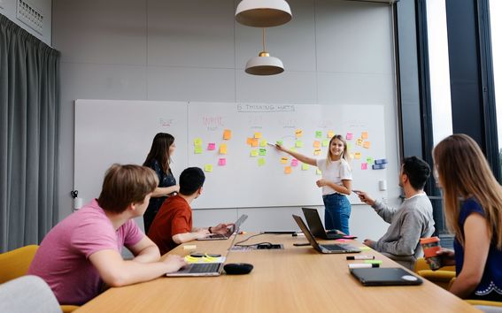 Pictured is a project room with a whiteboard and some post-it notes on the whiteboard. Students are sat and watching the teacher point at the whiteboard