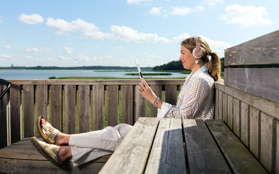 Henkilö istuu merinäköalan edessä kuulokkeet päässä ja selaa tablettia hymyillen.