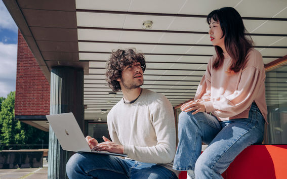 Two students sitting and discussing outside while one student is holding a laptop in his knee while listening to the other student.