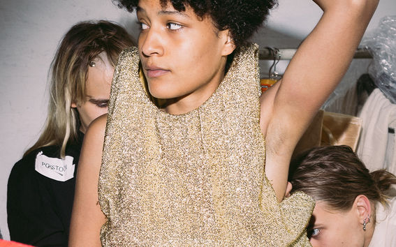 Malli odottamassa, kun kaksi ihmistä työstävät hänen päällään olevaa asua.