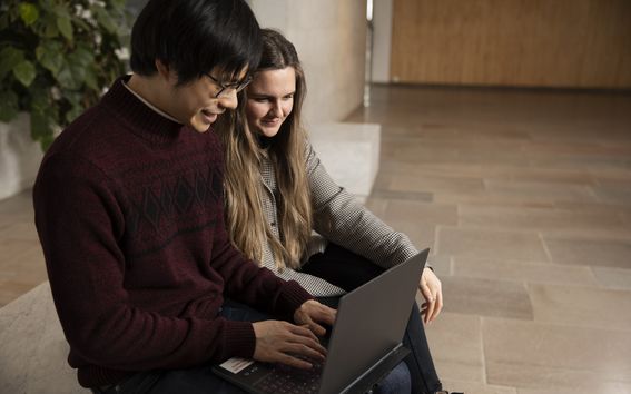 Two people are sitting on a bench in an empty hallway. One of them shows the other something from their laptop screen. 