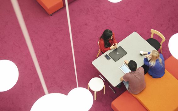 Three students photographed from above, students dressed in red and blue sitting at a table in a room with a pink floor