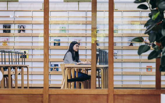 A student typing on a laptop pictured through a large glass wall in the library