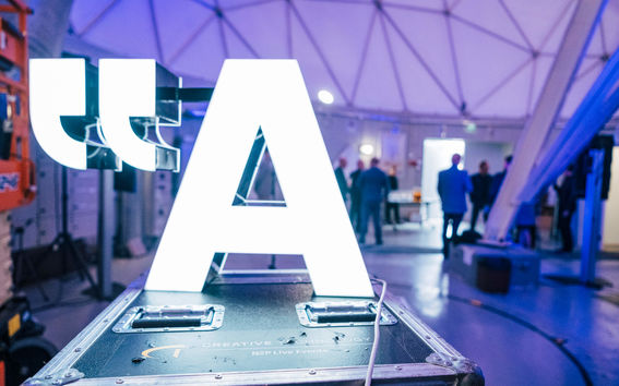 Aalto-yliopiston valaistu logo. Juhlavieraita logon takana tarkastelemassa radioantennia. 