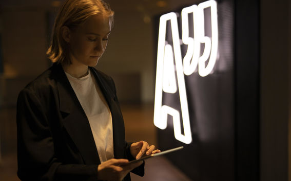 A student scrolling an ipad by an Aalto University logo neon sign.