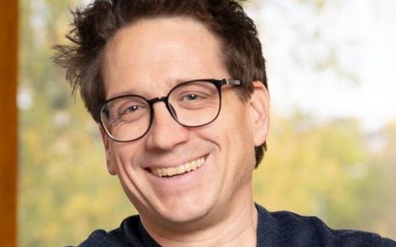 Marko Keskinen, jolla on lyhyet, tummat ja pörröiset hiukset, pukeutuneena tummansiniseen neuleeseen harmaan t-paidan päällä, istuu sisätiloissa.