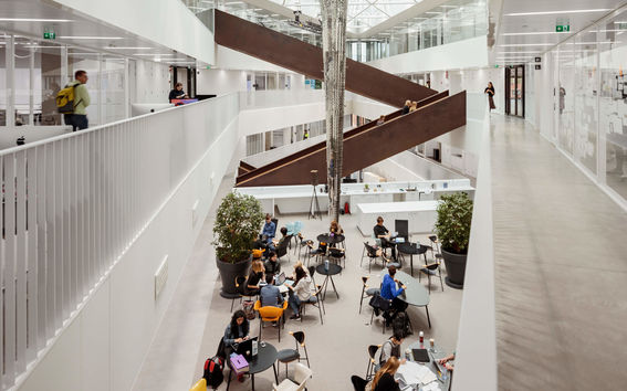 Modern flerplansatrium med takfönster, stor skulptur i mitten, studenter som arbetar vid bord och diagonal trappa.