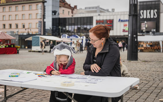 A child drawing outside at a table and an adult sitting next to her and watching her draw.
