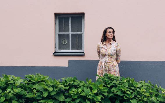 Kristjana Adalgeirsdottir photographed against a stone wall of a light pink house with a window in the background.