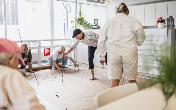 Five people drawing on brown paper on the ground with long black chalks.