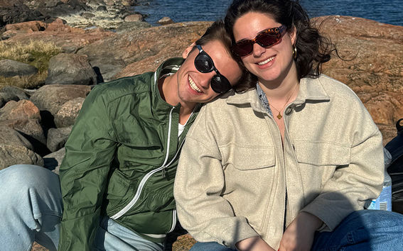 Two students hanging out on a beach in the spring, wearing green and beige jackets and sunglasses.