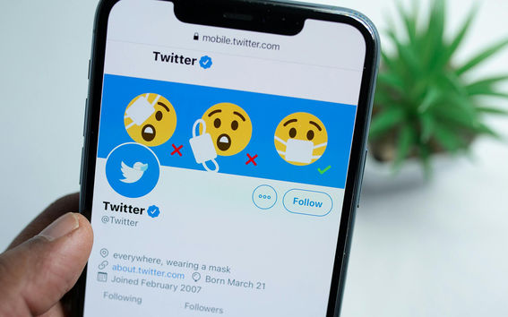 A person holds a phone showing the Twitter profile, with mask emojis in the header. A plant is in the background.