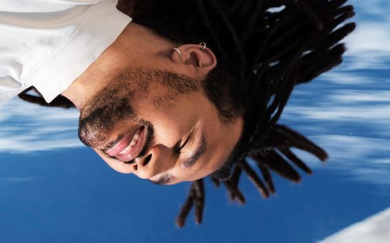 Person with dreadlocks and white shirt reflected in water against a blue sky with white clouds.