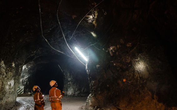 Two workers in orange protective gear and hard hats inspect a dimly lit rocky tunnel with overhead cables.
