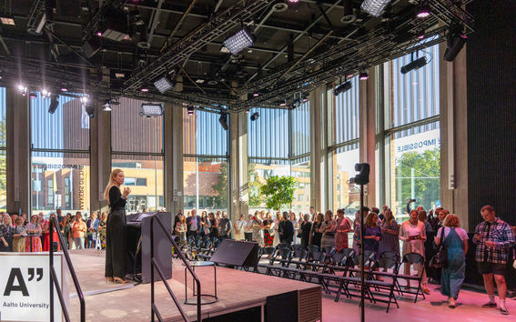 A speaker on a stage addressing a crowd in a modern building with large windows and high ceiling lighting.