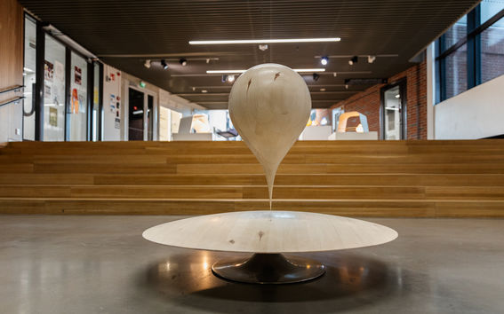 A wooden sculpture resembling a water droplet on a circular base, situated in a modern interior with wooden stairs.