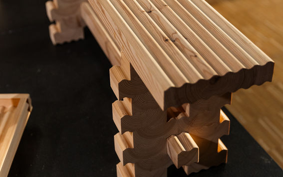 A wooden bench with a grooved surface and interlocking zigzag legs, placed in an indoor setting with wooden flooring.