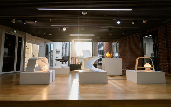A small exhibition room with unique chairs on white pedestals and various light fixtures, with large windows in the background.