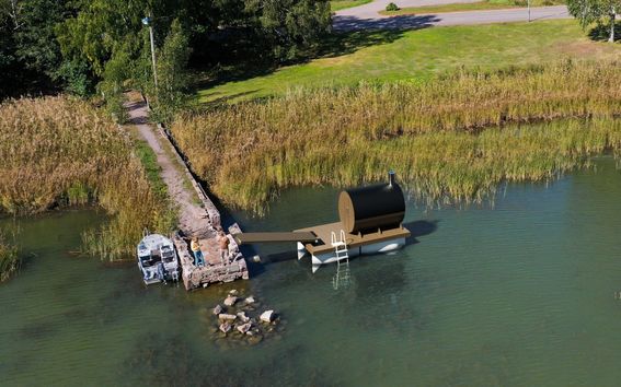 Ilmakuva laiturista, jossa kelluva sauna, pieni vene laiturilla ja kaislikkoinen ranta puineen.