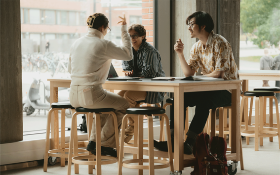 Tre personer sitter på pallar vid ett träbord i en modern inomhusmiljö, med suddig utsikt utanför.