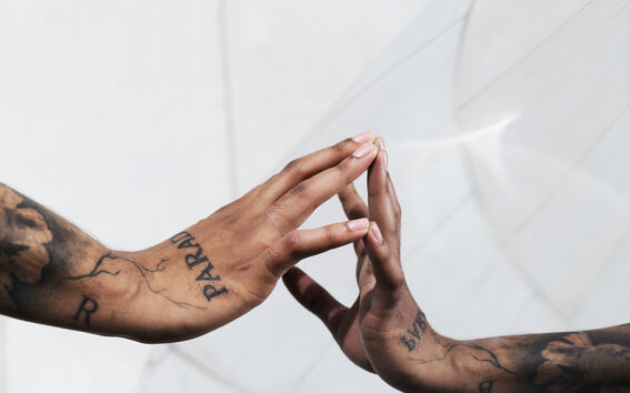 A tattooed arm with fingers touching its reflection on a glass surface, creating a mirrored effect.