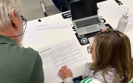 Two people working on experiment sheets at a table with a laptop and water bottle.