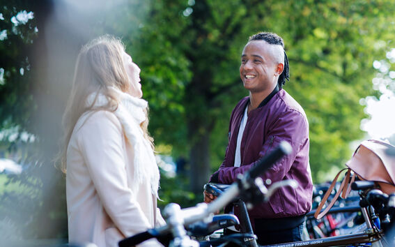 Kaksi ihmistä seisoo pysäköityjen polkupyörien lähellä, toinen valkoisessa takissa ja toinen violetissa takissa, ulkotilassa.