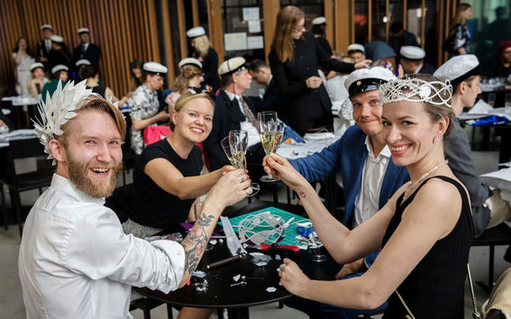People at a party are toasting with champagne glasses. Many wear hats and sit at tables with decorations.