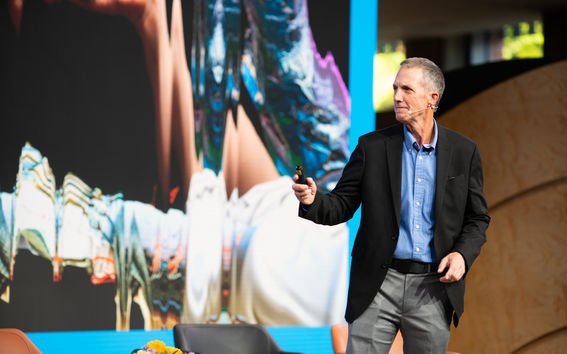 Mark Runco in a black blazer and blue shirt presents on stage with a blurred background image behind.
