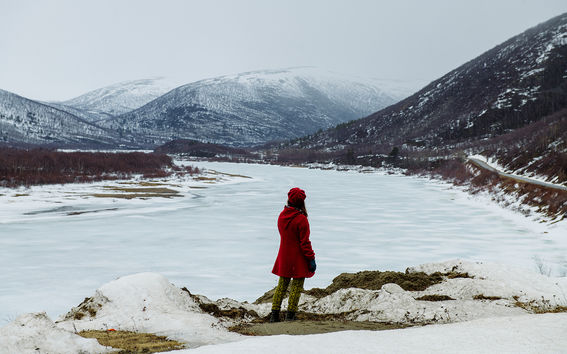 En person i röd kappa och hatt står på en snöhög och blickar över en frusen flod med berg i bakgrunden.