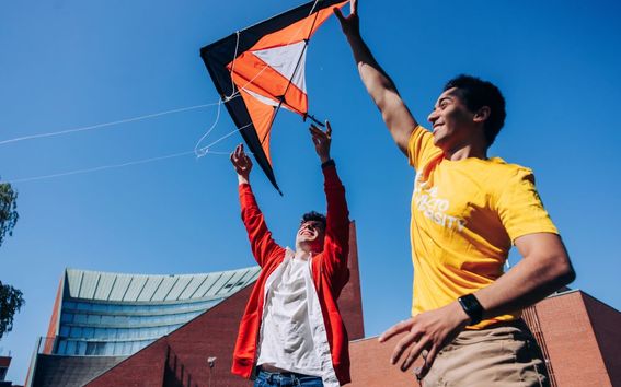 Two people fly a colourful kite outside with brick buildings in the background.