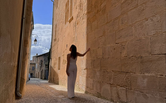Valkoisiin pukeutunut nainen seisoo kapeassa, aurinkoisessa kujalla, jossa on beigekiviseinät, koskien yhtä seinää ja katsoen ylös.