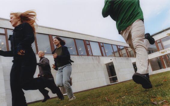 Four people running on a grassy area near a modern building with large windows.