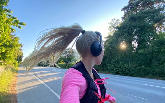 Henkilö, jolla on vaaleanpunainen paita lenkillä päällystetyllä tiellä, päässään kuulokkeet ja liivi, taustalla puita ja pensaita.