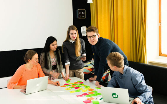 A group of people collaborate around a table covered with sticky notes and laptops in a modern office.