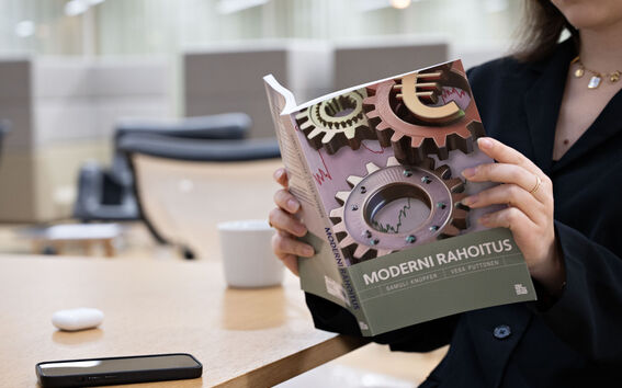 Person reading a book titled 'Moderni Rahoitus' at a table with a smartphone and a coffee cup.