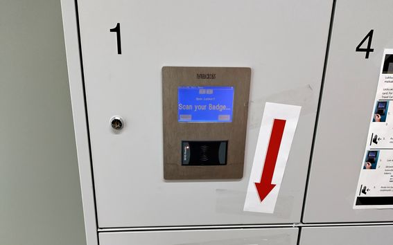 Locker system with a digital display reading 'Scan your Badge'. A large red arrow points down next to the screen.