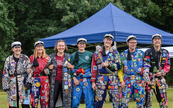 A group of people in colourful student overalls with patches standing arm in arm in front of a blue tent in a grassy area.