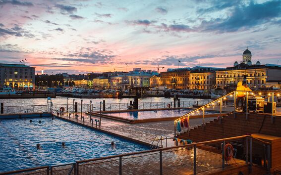 Evening view of a pool by a harbour with illuminated buildings and a church in the background.