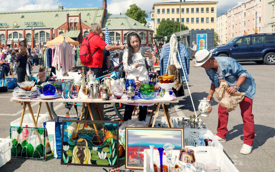 Outdoor flea market with tables displaying glassware, ceramics, and framed art. People browsing items under a sunny sky.