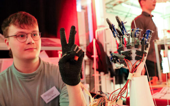 Hanskaa käyttävä henkilö näyttää rauhanmerkkiä mekaanisen käden vieressä, jossa on johtoja ja antureita.