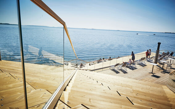 People relax on wooden terraces leading to the sea on a sunny day, with a glass railing on the left side.