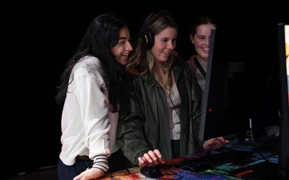 Kolme henkilöä on kokoontunut tietokoneen näytön ääreen, yksi heistä käyttää kuulokkeita ja hiirtä.