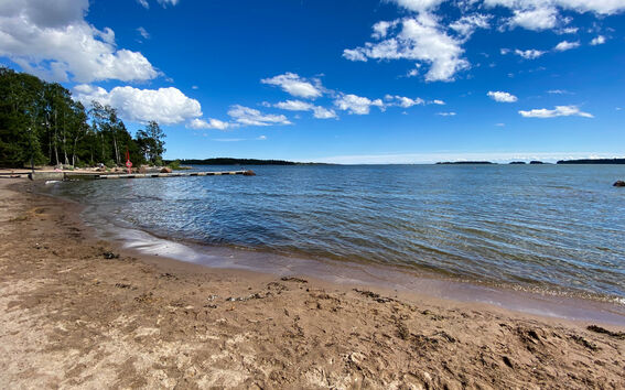 A sandy beach by a calm sea with a pier and trees in the background under a partly cloudy sky.