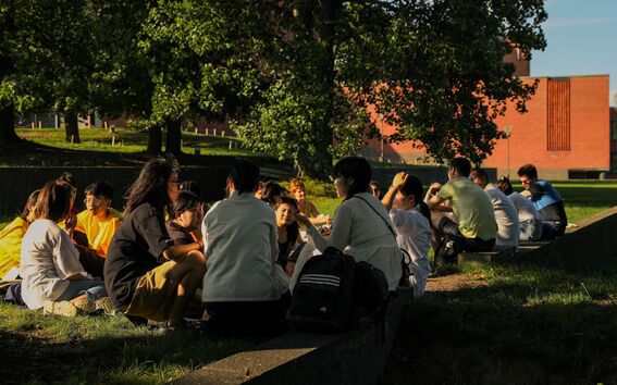 A group of people sitting on the grass in a park, with trees in the background and a red brick building behind them.