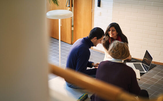 Tre personer sitter på golvet i en korridor med en bärbar dator och diskuterar.
